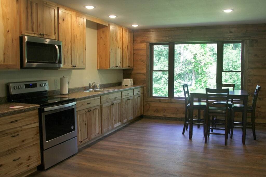 A full kitchen with wooden cabinetry and modern appliances is showcased. A dining table with four chairs is positioned near a large window that overlooks the surrounding greenery. Soft, natural light enhances the warm tones of the wood and flooring.
