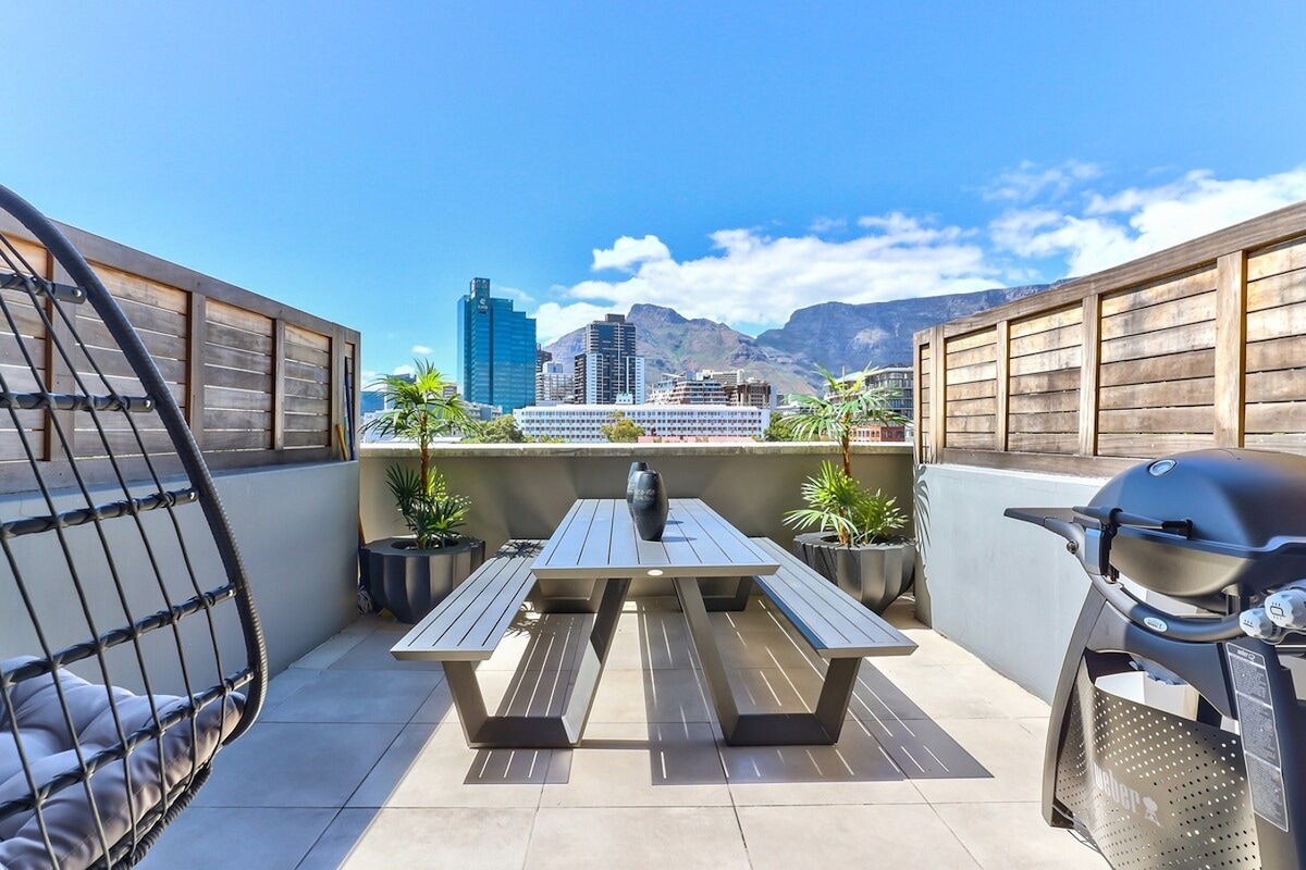 An outdoor terrace is showcased with a dining table surrounded by potted plants. A charcoal grill is positioned near the edge. Views of Table Mountain and the city skyline are visible in the background under a clear blue sky.