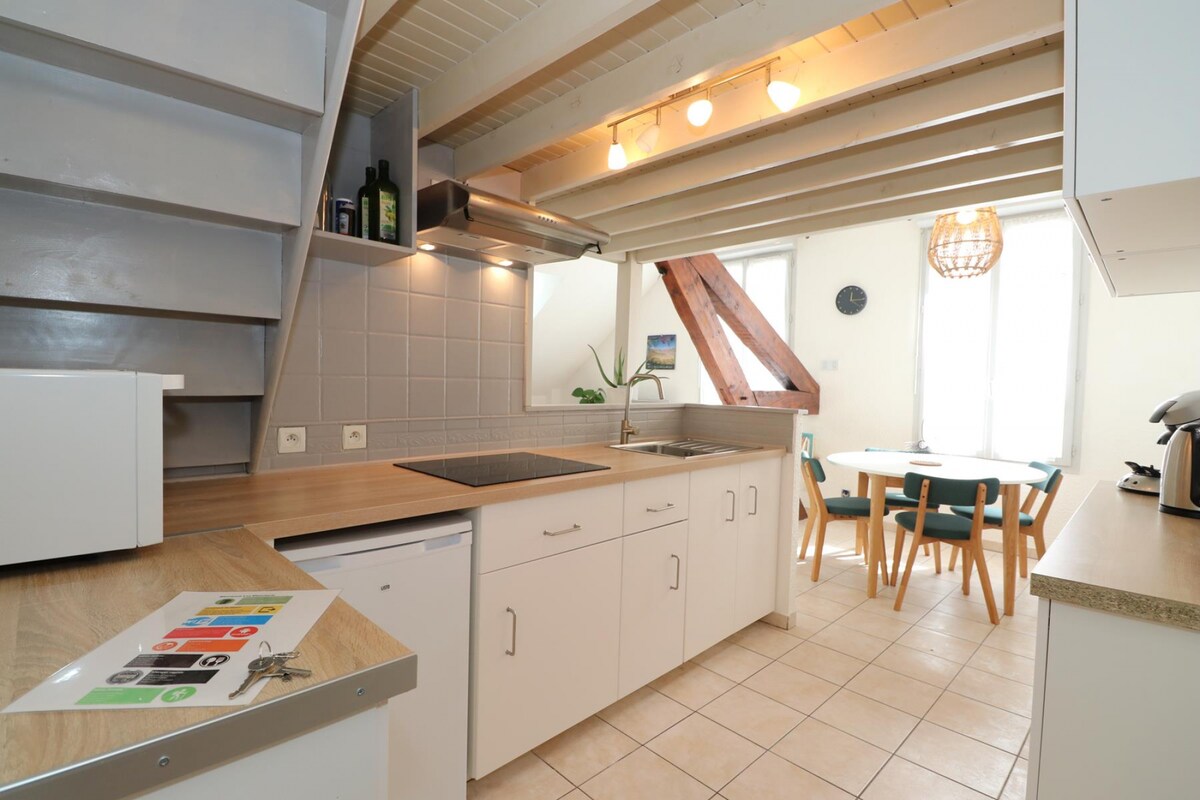 A well-equipped kitchen features modern appliances and a spacious countertop. Natural light streams through windows, highlighting the wooden beams above. A dining area with a circular table and four teal chairs is visible, creating a functional and inviting space.