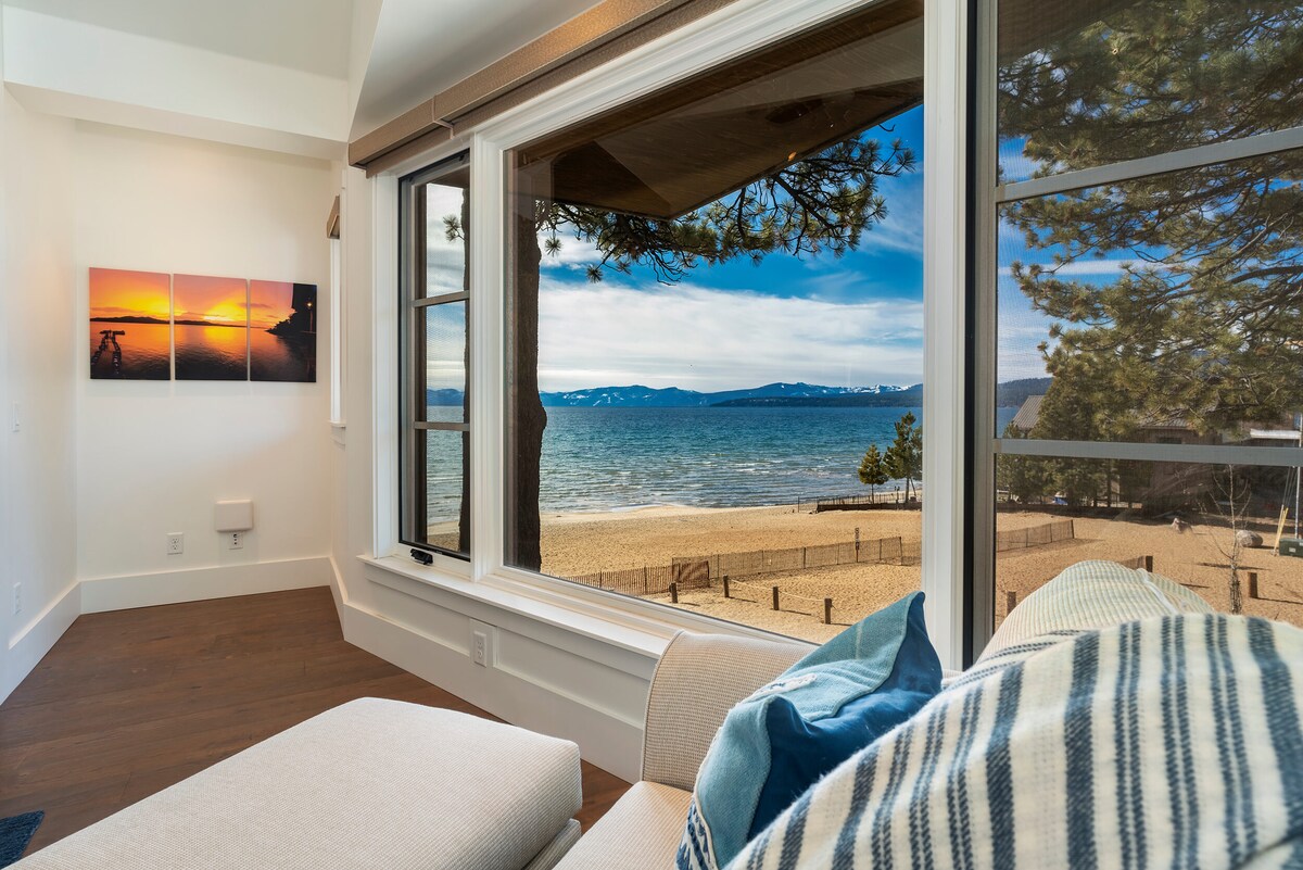 A comfortable living area is framed by expansive windows showcasing views of Lake Tahoe and a sandy shoreline. The soft furnishings, including a large sectional couch with decorative pillows, invite relaxation as the setting sun paints the sky in warm hues.