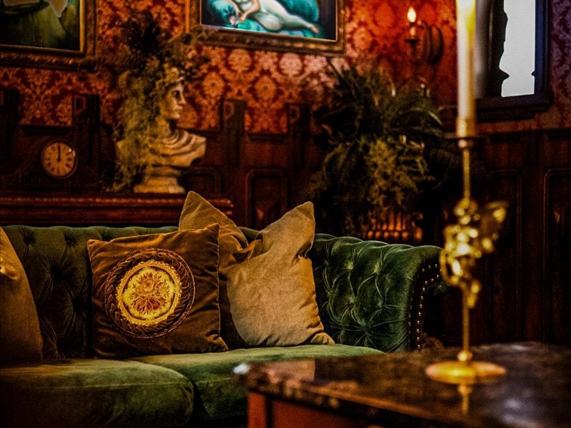 A rich green velvet couch is adorned with decorative pillows featuring intricate designs. The surrounding walls are decorated with ornate wallpaper and various artworks, while a softly glowing candle adds a touch of warmth to the inviting space.