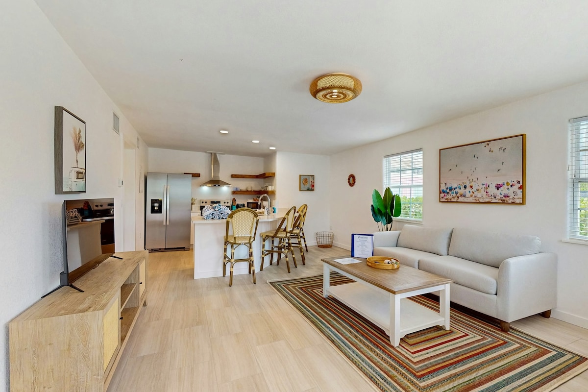 A spacious living area is equipped with a light-colored sofa and an area rug featuring muted stripes. A dining table with rattan chairs is visible in the background, adjacent to a fully-equipped kitchen with stainless-steel appliances. Natural light filters in through multiple windows.