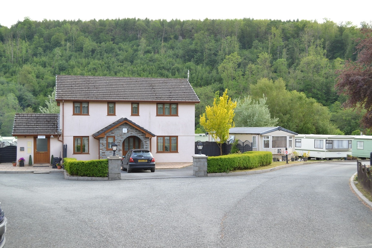 A two-story building with a light pink exterior is shown in a peaceful wooded setting. A grey car is parked in front of the main entrance, while several caravans are visible along the curved path. Lush greenery surrounds the area, complementing the natural backdrop.