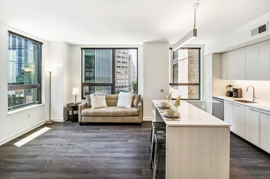 A modern living space features large windows allowing abundant natural light. A cozy couch sits beside a floor lamp, with a sleek kitchen area visible in the background. The open layout is complemented by hardwood floors and a long countertop with dining stools.