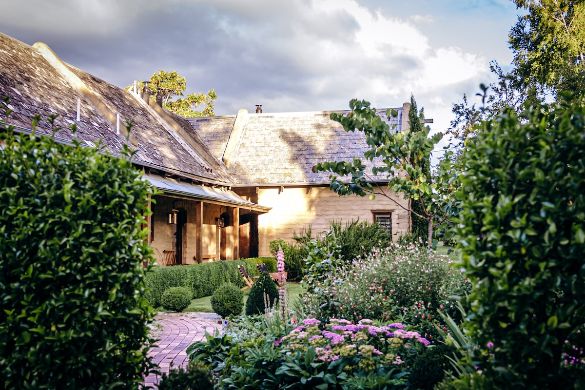 The exterior of the Stable Lofts is framed by neatly trimmed hedges and a vibrant garden filled with blooming flowers. The rustic mudbrick structure features a charming patio area that invites relaxation, all set against a backdrop of a partly cloudy sky.