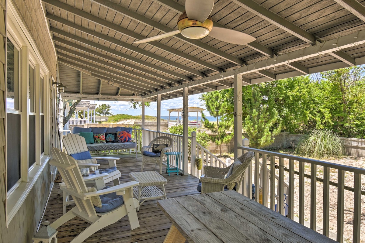 A spacious covered porch features a wooden floor and ceiling fan, providing a relaxed outdoor space. Multiple seating options include adirondack chairs and a low table. The porch overlooks a private yard with greenery and offers views of the nearby beach.