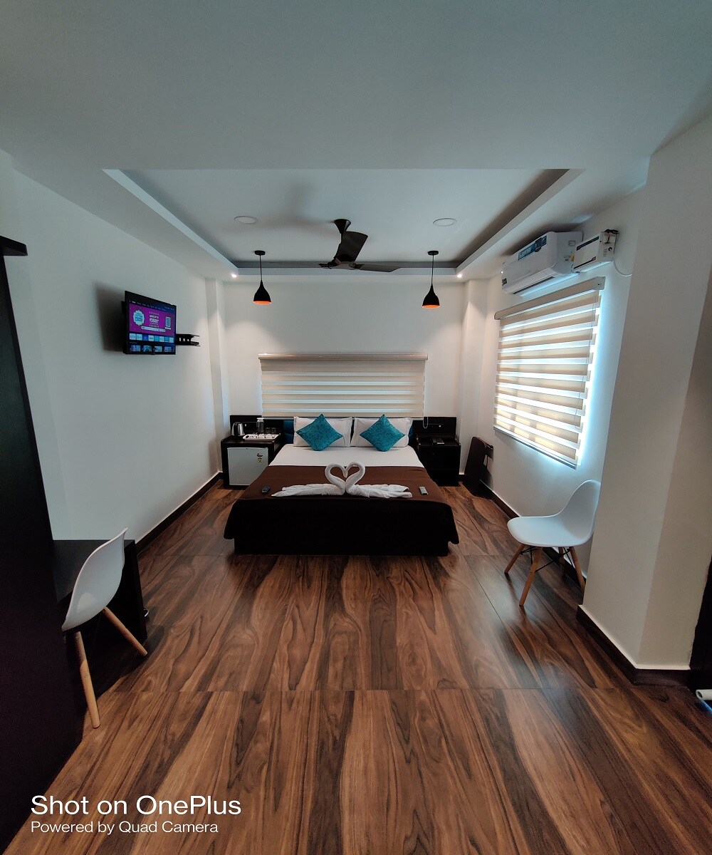 A modern room features a spacious bed adorned with decorative towels shaped like swans. Light-colored walls are complemented by wooden flooring. Two white chairs are positioned near a compact table. A flat-screen TV is mounted on the wall, and large windows provide natural light.