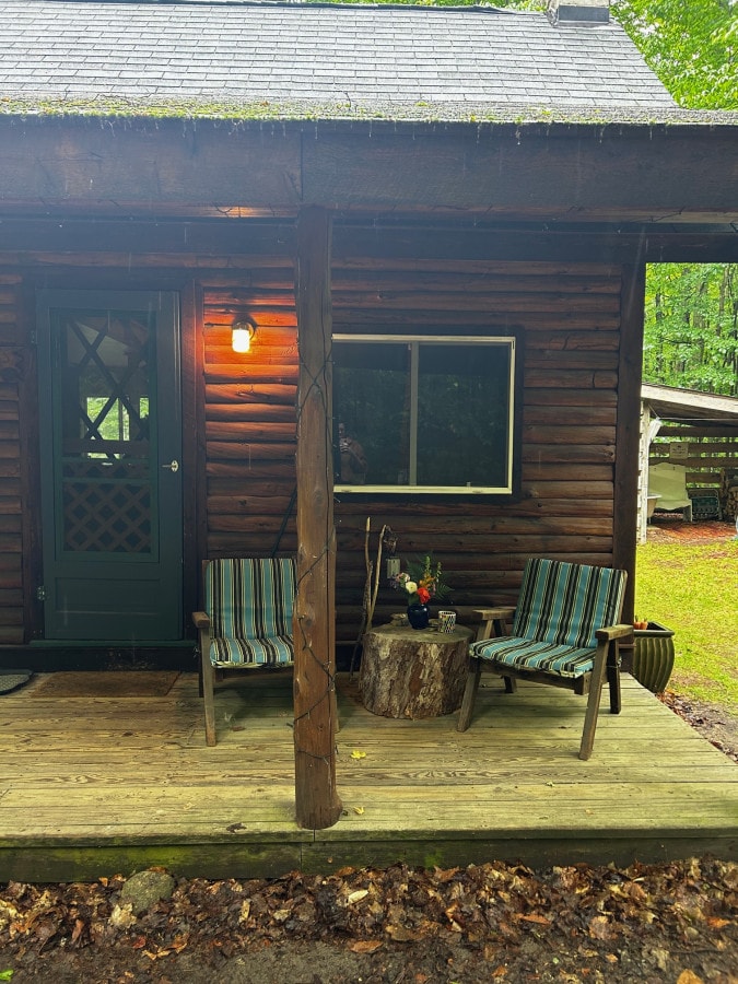 A wooden porch features two striped chairs positioned on either side of a small tree stump table. A potted plant and a flower arrangement are placed on the stump, creating a welcoming entrance to the cabin. Soft lighting illuminates the area, enhancing the natural surroundings.