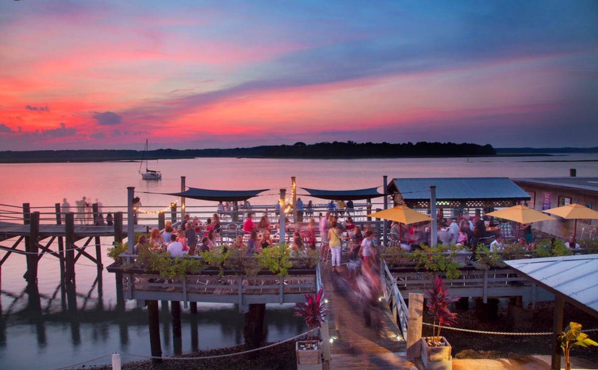 A vibrant waterfront restaurant is depicted at sunset, showcasing outdoor seating under umbrellas. The boardwalk leads towards the restaurant, with lively diners visible against the backdrop of a colorful sky reflecting on the calm water, creating a serene dining atmosphere.