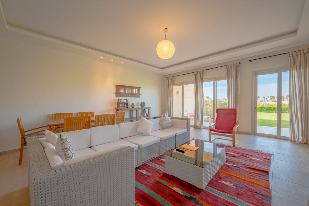 A spacious living area features a white sectional sofa paired with a red accent chair. A vibrant area rug adds color to the space. Large sliding glass doors provide views of the garden, while a wooden dining table is positioned in the background.