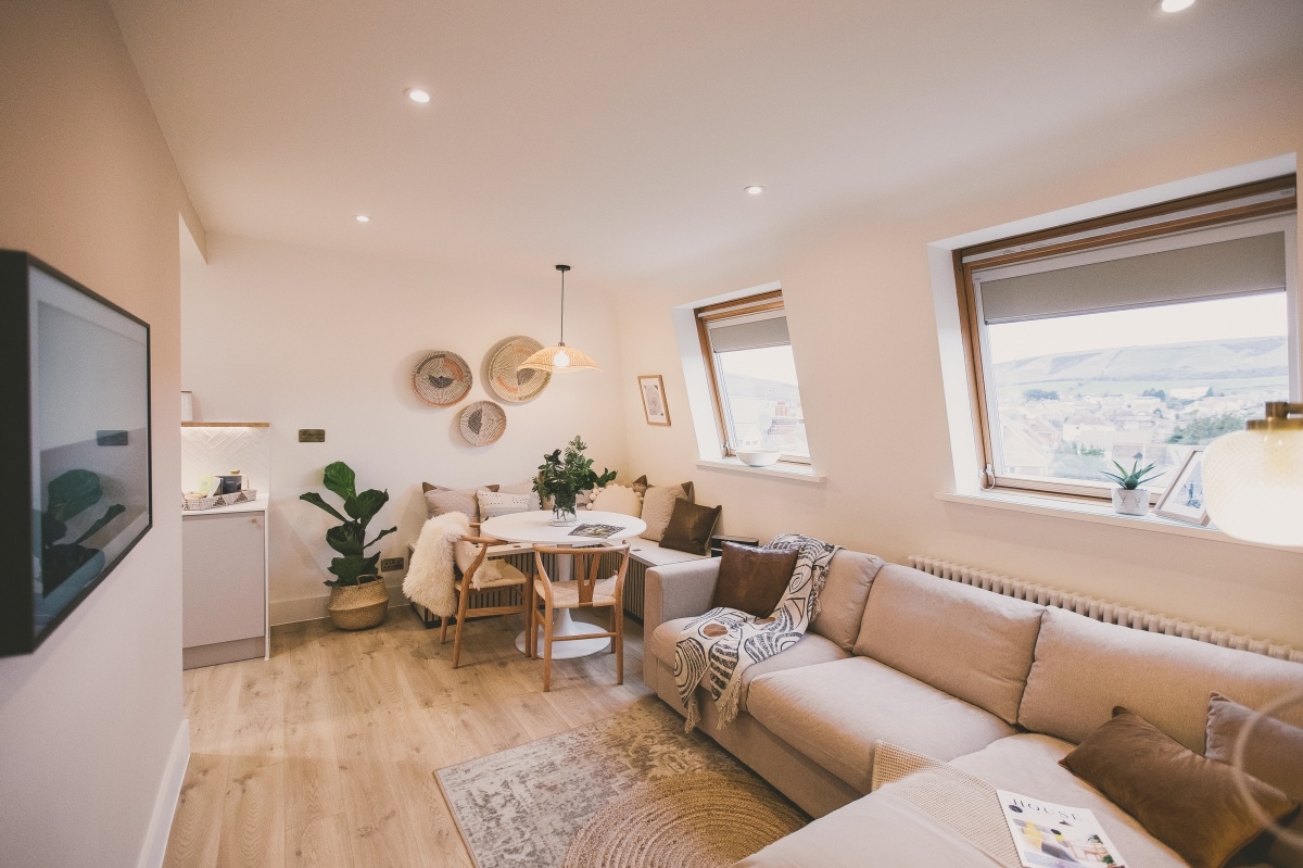 A modern living space features a light-colored sectional sofa with decorative cushions and a round dining table surrounded by chairs. Two windows invite natural light, while a plant adds a touch of greenery. A wall-mounted TV complements the contemporary decor.