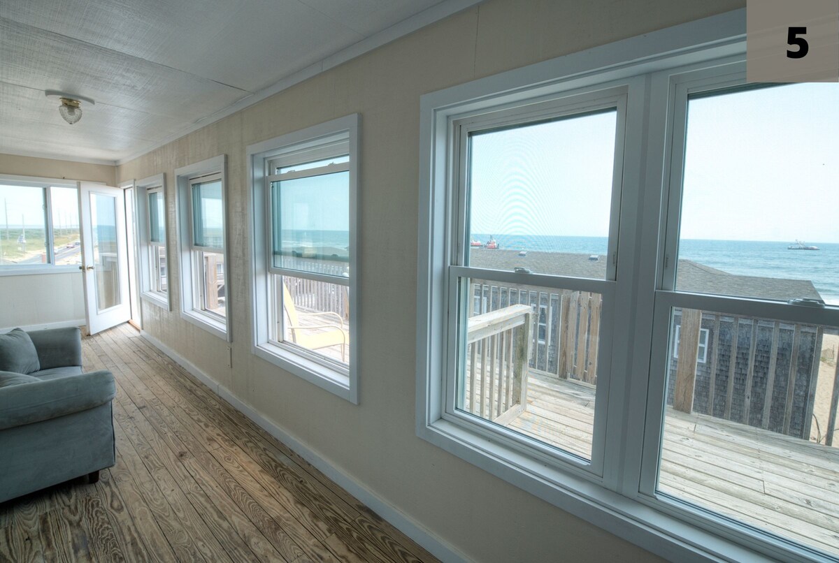 Large windows allow natural light to fill the room, providing scenic views of the beach and ocean. Wooden flooring complements the neutral walls, and a comfortable sofa is positioned nearby. A spacious outdoor deck is visible through the windows, enhancing the coastal atmosphere.