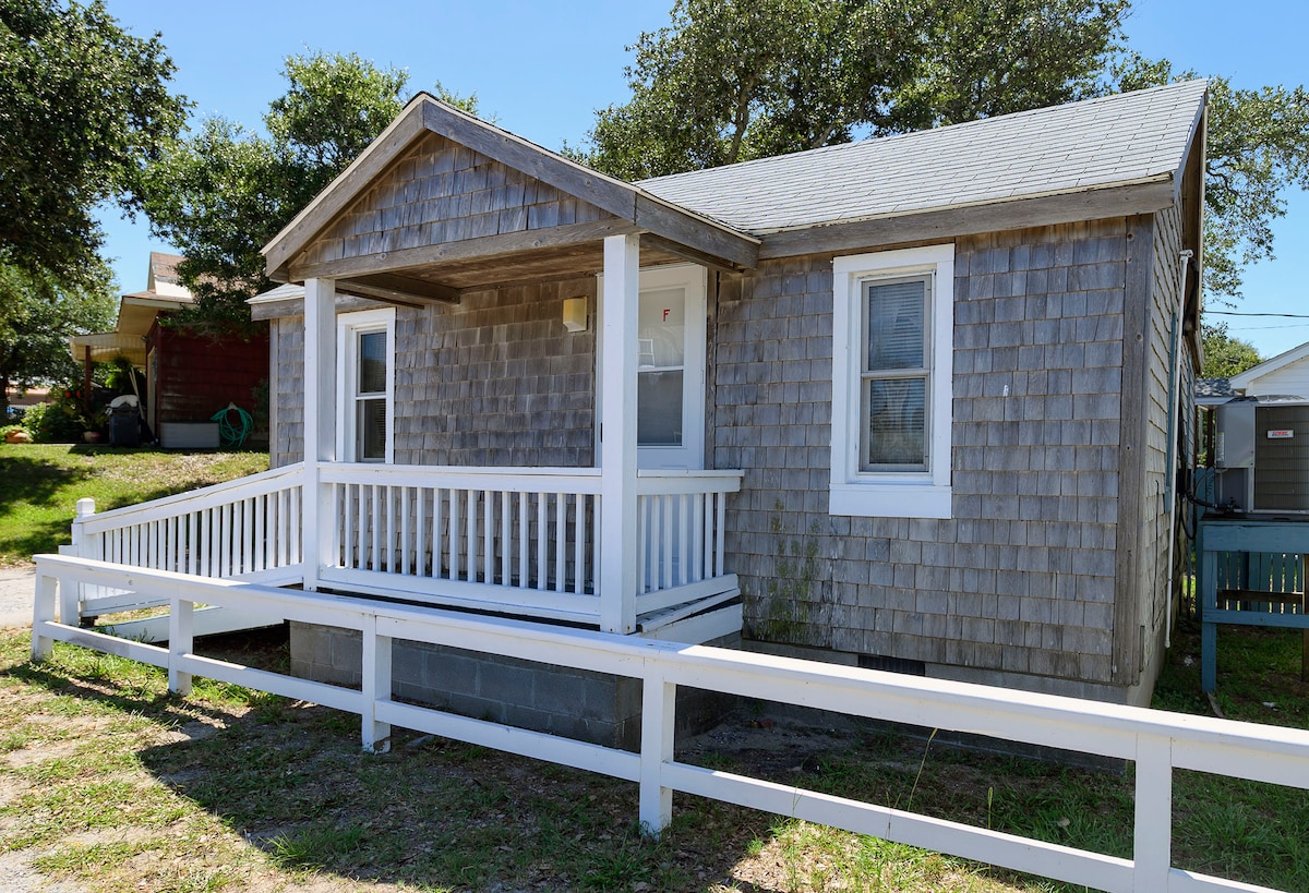 A single-story cottage is shown with a weathered shingle exterior, featuring a covered front porch supported by clean white railings. Two windows on the front allow natural light to enter, while lush greenery surrounds the property, enhancing the outdoor setting.