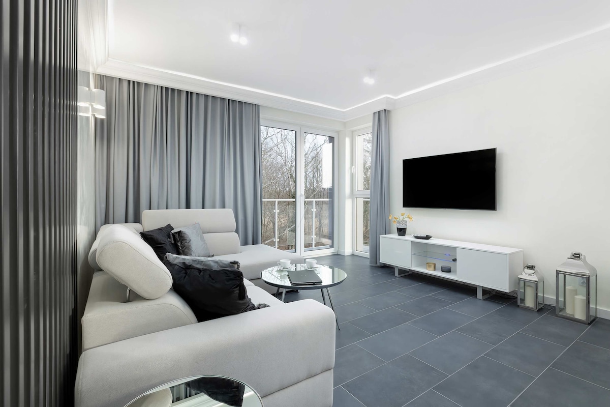 A modern living room features a light gray sofa with black throw pillows. A glass coffee table sits in the center, and a wall-mounted TV is positioned opposite. Large windows with sheer curtains provide natural light, while a balcony can be accessed from this space.