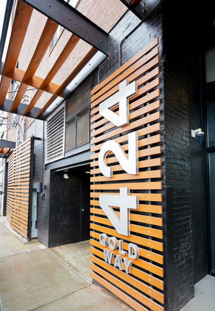 The entrance of the building is highlighted by wooden slats and contemporary signage displaying '424 GOLD WAY.' The façade features a mix of brick and concrete, reflecting an industrial modern design. The pathway is paved, leading towards the entrance.