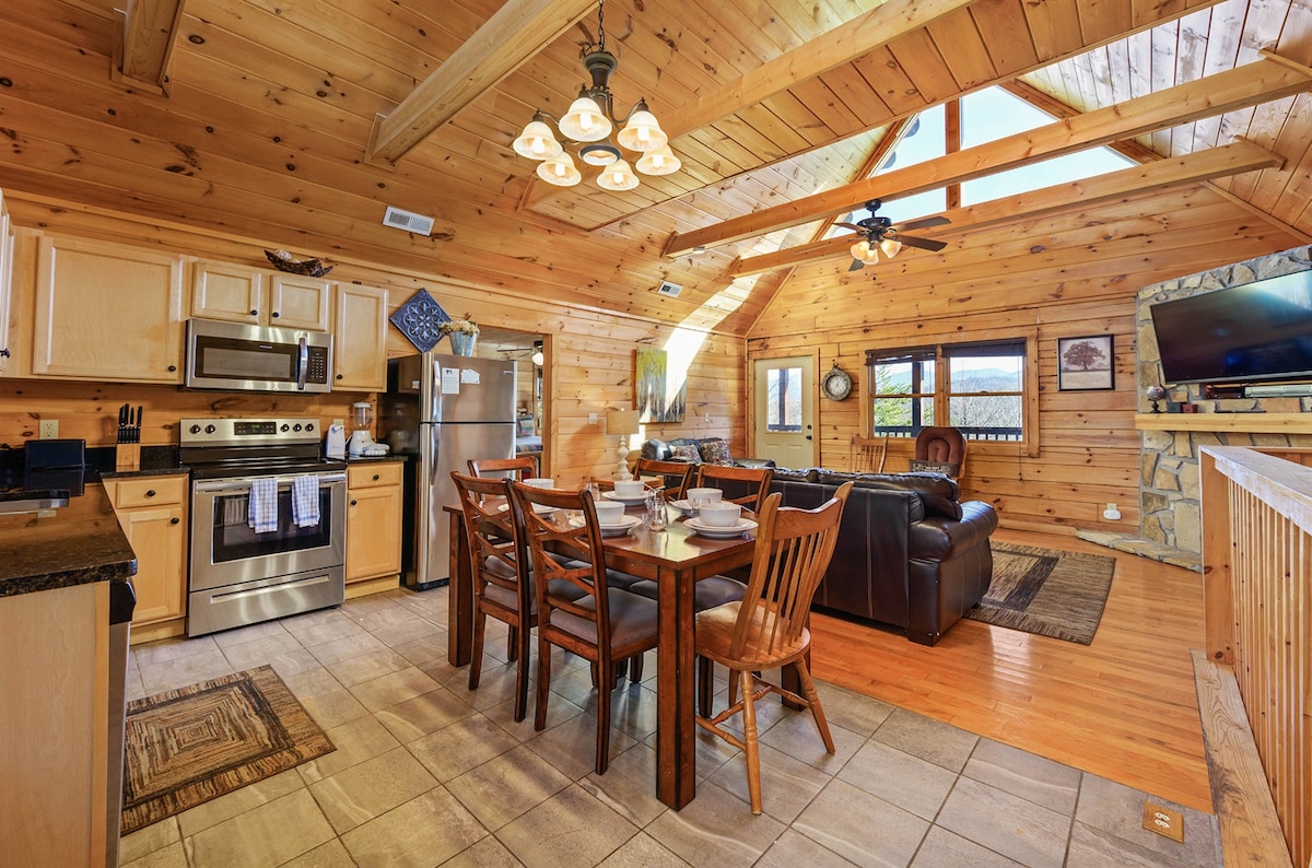 An open-concept living space features a warm wooden interior with vaulted ceilings. A dining table for six is positioned near the kitchen, where stainless steel appliances are visible. A comfortable seating area includes leather couches, and large windows enhance the natural light and views.
