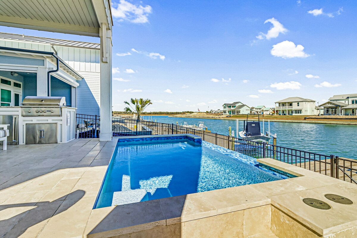 A private pool overlooks the canal, with clear blue water reflecting the sky. A large outdoor kitchen is positioned nearby, featuring a built-in grill. The surrounding deck is bordered by a low fence, offering unobstructed views of the water and nearby homes.