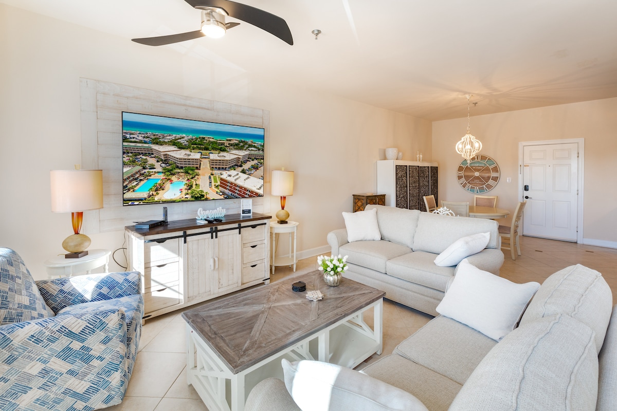 The living area features a large flat-screen television mounted above a stylish media console. Comfortable seating includes a light-colored sofa and patterned armchairs. A wooden coffee table sits in the center, adorned with a vase of flowers. Natural light fills the room from windows and a ceiling fan hangs above.
