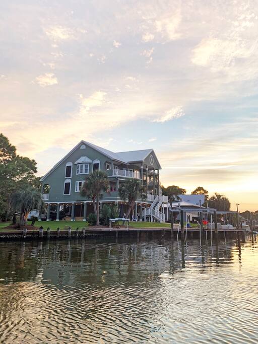 The three-story home features a blend of modern and coastal architecture, highlighted by large windows and a wraparound porch. Set against a colorful sunset, its landscaped yard and palm trees can be seen bordering the reflective waters of the cove.