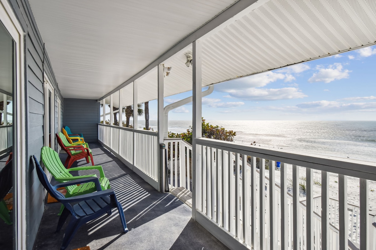 A covered balcony features colorful chairs in green and blue, offering a comfortable space for relaxation. The ocean can be seen in the distance, with sunlight reflecting on the water, enhancing the coastal atmosphere.