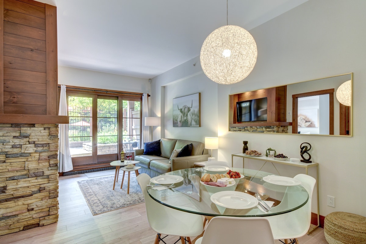 The dining area features a round glass table set with plates, accompanied by a light fixture overhead. A cozy seating area with a sofa is visible in the background, along with large windows that provide access to an outdoor space. A neutral color palette enhances the inviting ambiance.