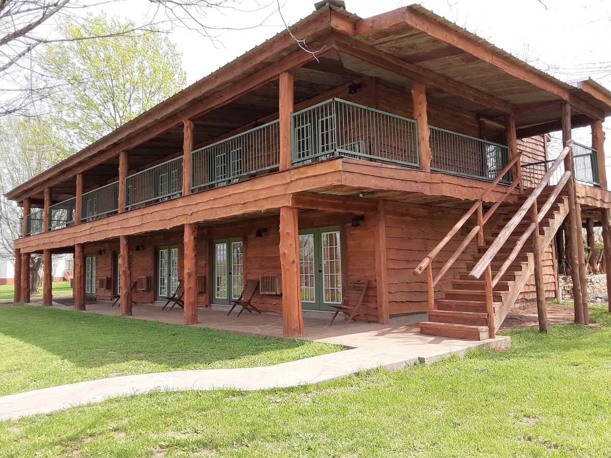 A wooden lodge with a spacious porch features multiple sets of French doors, providing access to the guest rooms below. Sturdy stairs lead to the upper level. Lush green grass surrounds the building, enhancing the natural setting.