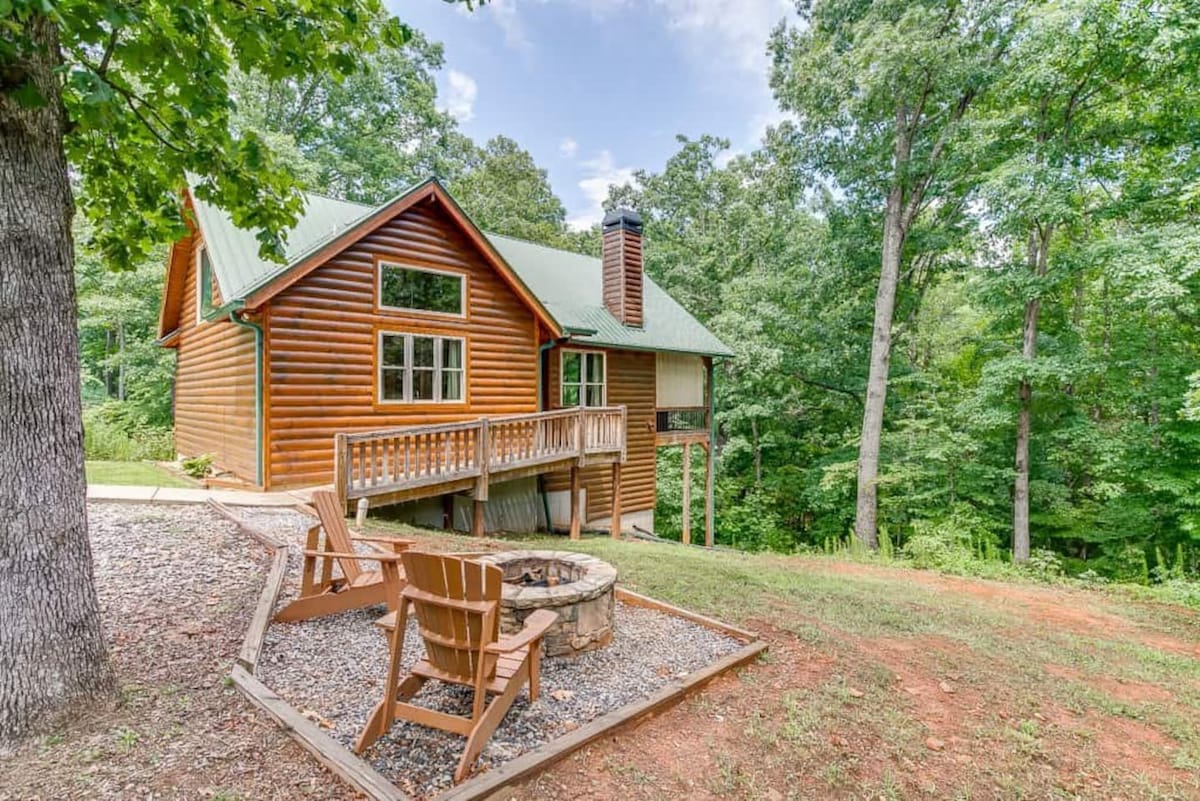 A rustic cabin exterior is framed by lush green trees, with a wooden deck extending from the side. A stone fire pit sits surrounded by gravel and wooden seating, inviting cozy gatherings in a serene woodland setting.