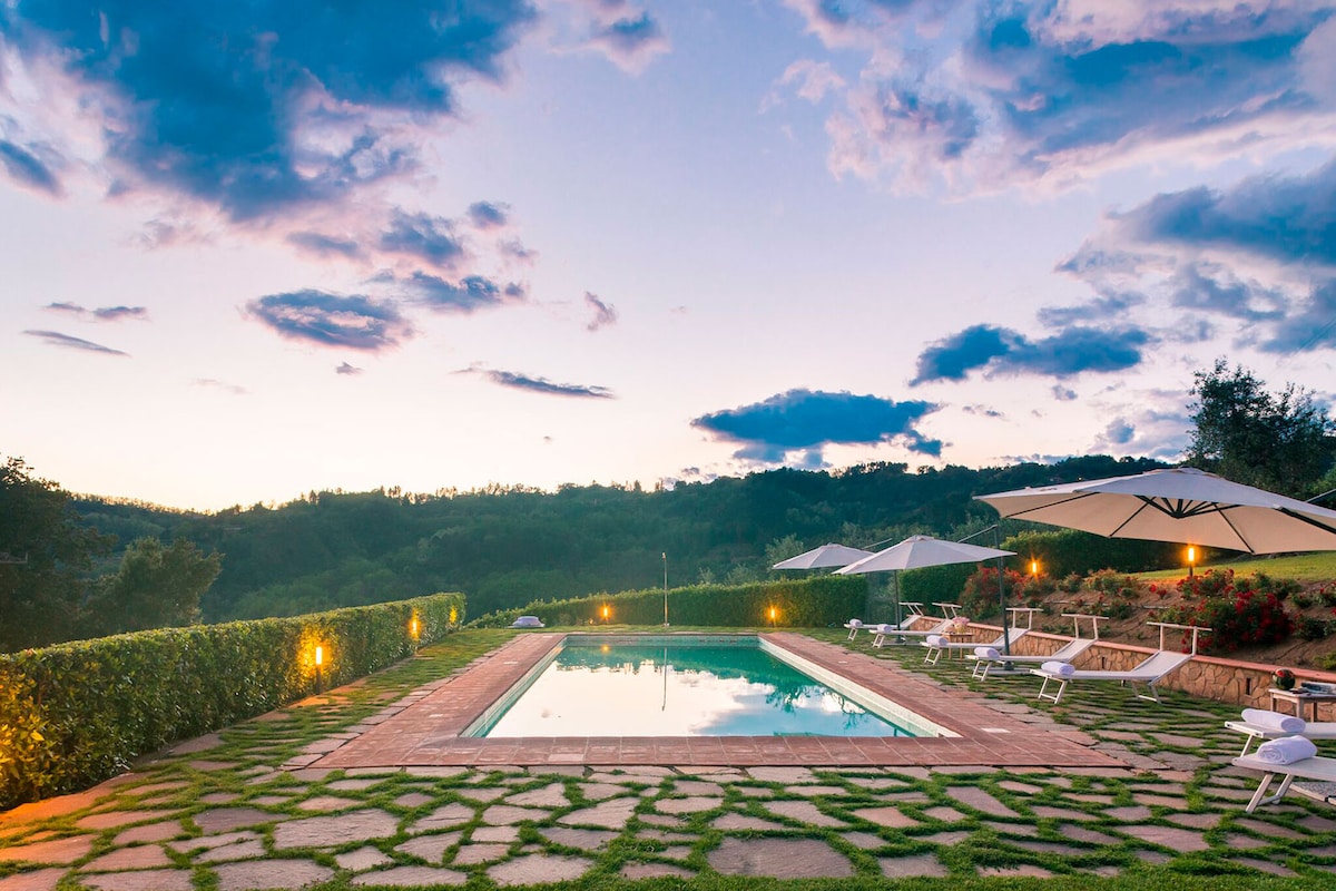 An inviting pool area is surrounded by lush greenery and stone paths. Comfortable lounge chairs and large umbrellas are positioned around the pool, which reflects the evening sky. Soft lighting enhances the serene atmosphere as dusk approaches, offering a peaceful outdoor retreat.