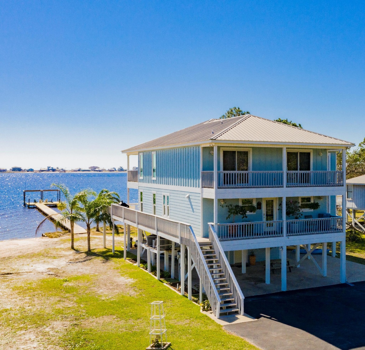 An expansive beach house is situated beside a calm lagoon, featuring multiple levels and a private pier. Lush greenery surrounds the base of the house, while steps lead up to the wraparound decks. Clear skies reflect off the water, enhancing the serene setting.