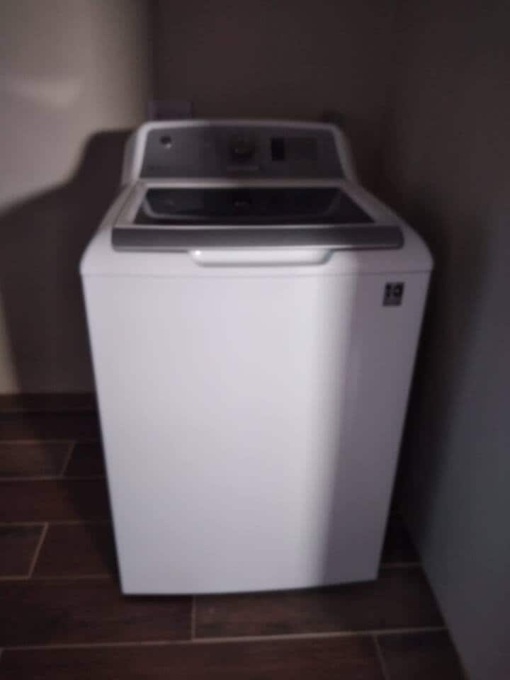 A top-loading washing machine is positioned in a corner, featuring a sleek white exterior. The machine is equipped with a control panel on the back, and the surrounding area showcases tiled flooring that complements the appliance's modern design.