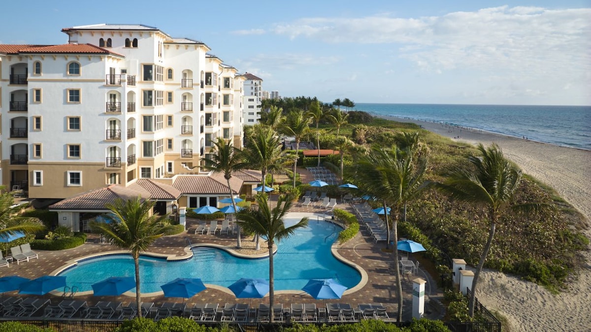 The resort's vibrant pool area is framed by palm trees, with blue umbrellas offering shade. The spacious pool, set against the backdrop of the beachfront, is accompanied by lounge chairs. The ocean and beach stretch out in the distance under a clear sky.