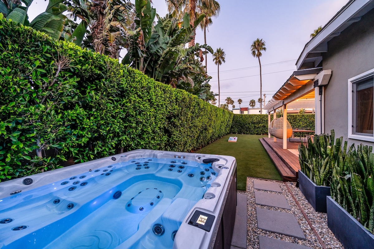 A spacious backyard is highlighted by a 10-person hot tub, surrounded by well-maintained hedges for privacy. Lush greenery and palm trees create a serene atmosphere, with a grassy area and a horseshoe pit visible in the background. Soft lighting enhances the tranquil setting.