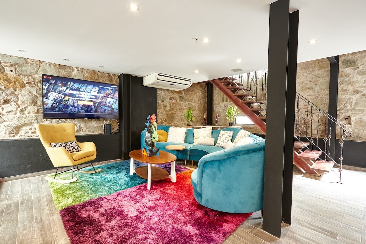 A spacious living area is highlighted by a vibrant rug displaying a gradient of colors. A modern blue sofa and a yellow armchair offer seating options. A large Smart TV is mounted on the wall, and a wooden table is placed at the center. A staircase leads to the upper level.