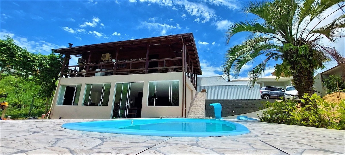 A modern house with a welcoming design is showcased, featuring a private pool with a light blue edge. Large glass windows reflect the surrounding greenery, while a wooden balcony extends over the structure. The sky is bright with scattered clouds, enhancing the peaceful atmosphere.