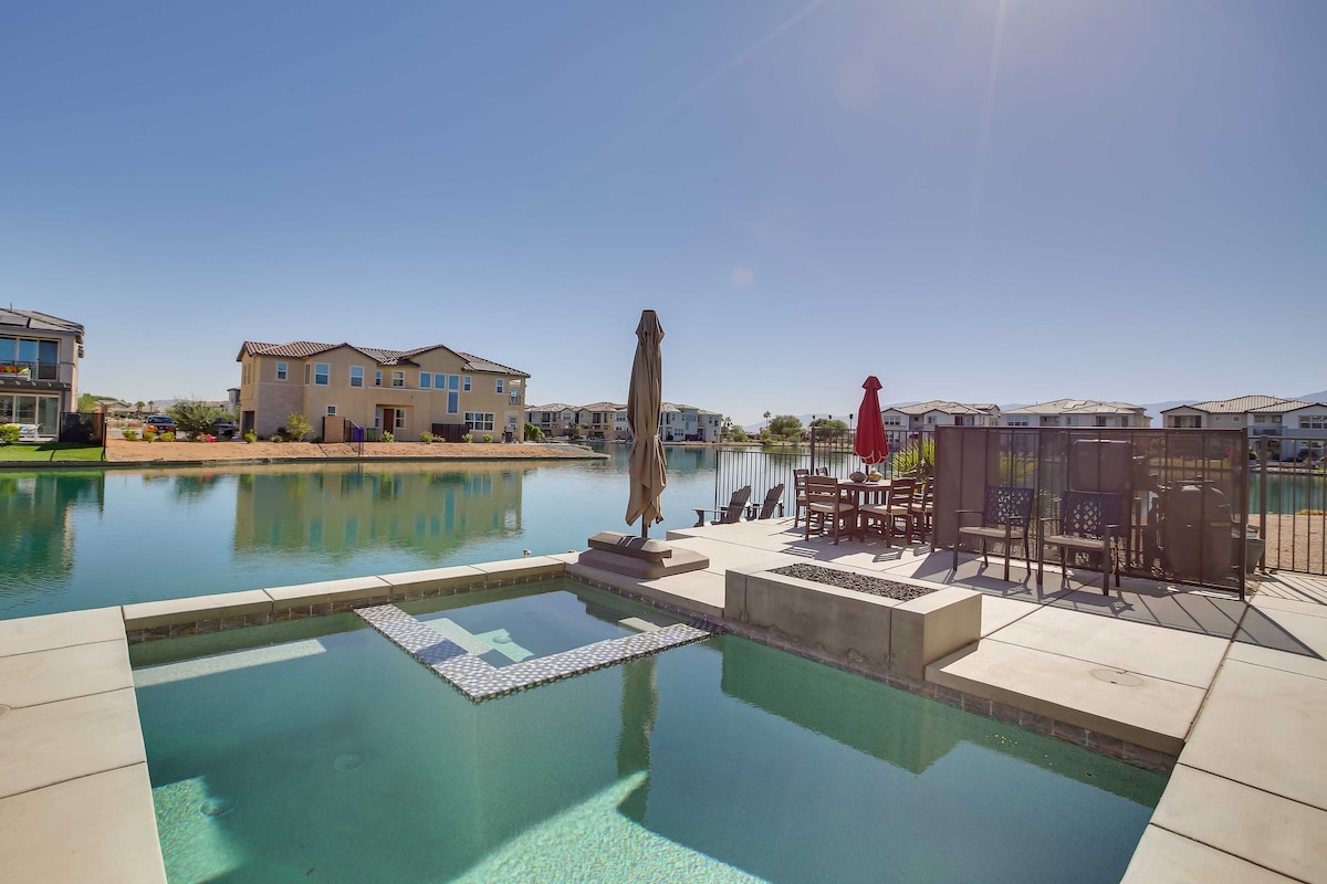 A serene outdoor space features a heated saltwater pool and a hot tub, surrounded by lounge chairs and a dining area with umbrellas. Reflections of nearby homes and clear blue skies enhance the tranquil atmosphere of the water view.