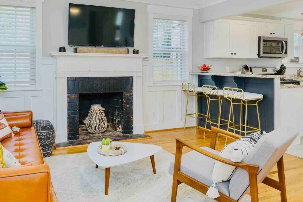 A living area features a leather sofa paired with an accent chair, set on a light rug. A modern coffee table is centered, while a TV is mounted above a classic fireplace. The kitchen bar with golden stools is visible in the background, offering additional seating.