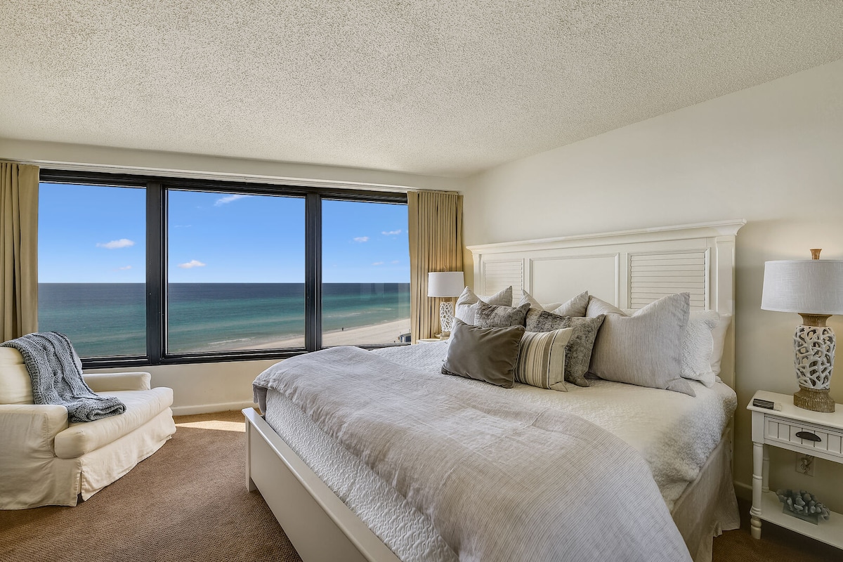 A serene bedroom is highlighted by a spacious bed dressed in light, neutral linens. Large windows offer sweeping views of the ocean and beach, allowing natural light to fill the room. A cozy armchair and bedside tables with lamps add a touch of comfort.