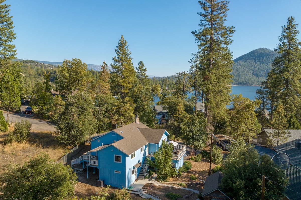 A two-story blue house is surrounded by tall trees and greenery, offering a scenic view of the nearby lake and mountains. The property features a welcoming outdoor space and is situated within a tranquil neighborhood, with pathways and roads visible in the surrounding area.