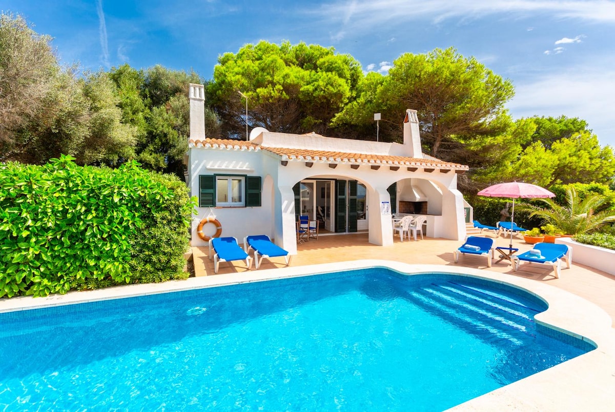 The exterior of the villa features a private pool surrounded by sun loungers. A covered terrace is visible, leading into the living area. Lush greenery and trees provide shade, while a pink umbrella adds a touch of color to the outdoor space.