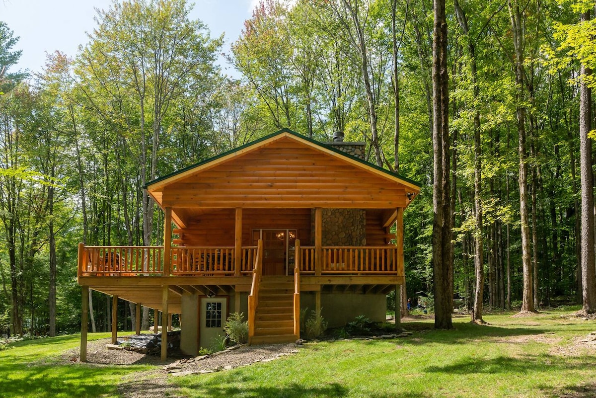A spacious log cabin is nestled among lush trees, featuring a large wrap-around deck with wooden railings. Steps lead up to the entrance, welcoming visitors to explore the inviting nature of the surroundings and enjoy the tranquility of the forest.