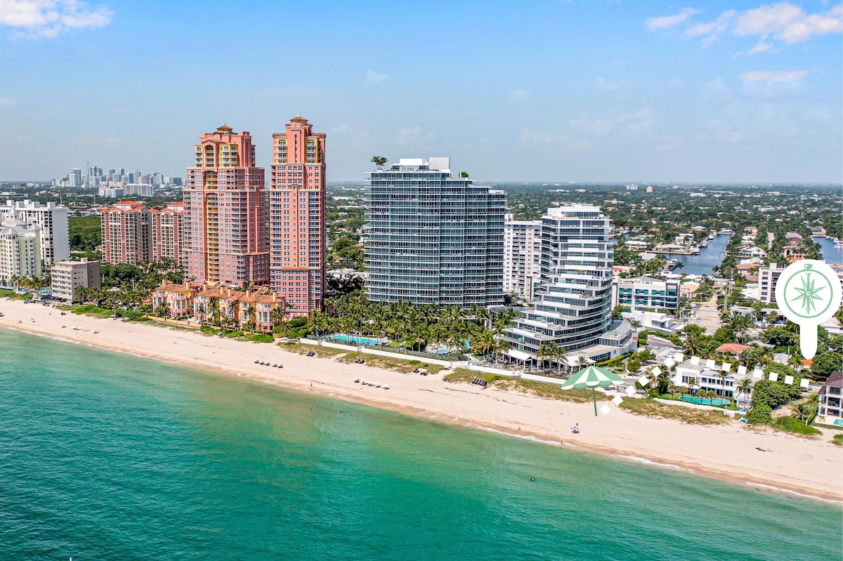An aerial view captures the coastline lined with sandy beaches and upscale buildings, showcasing a vibrant mix of high-rise structures and tropical foliage. The calm turquoise waters reflect the sunny sky, while the beach invites relaxation and leisure activities against a backdrop of the city skyline.