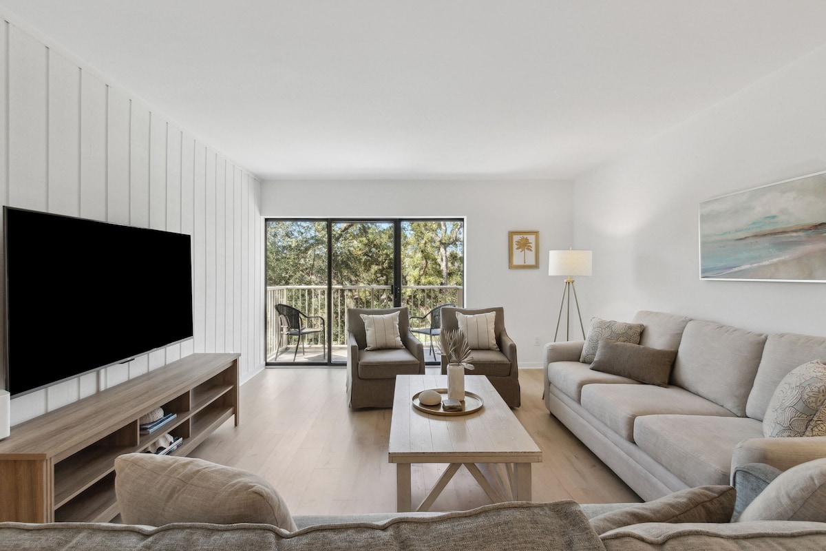 A light-filled living area is depicted, featuring a comfortable gray sectional sofa and armchairs, with a natural wood coffee table positioned centrally. Large glass doors open to a balcony, offering views of the surrounding greenery. A modern television is mounted on the wall opposite the seating area.