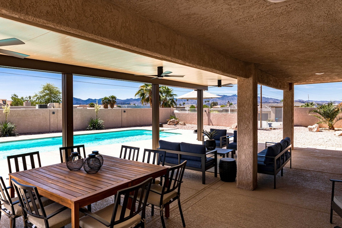 An outdoor patio area features a wooden dining table surrounded by black chairs, offering a space for meals. A private pool is visible in the background, framed by desert landscaping and mountain views. Ceiling fans are installed for airflow.