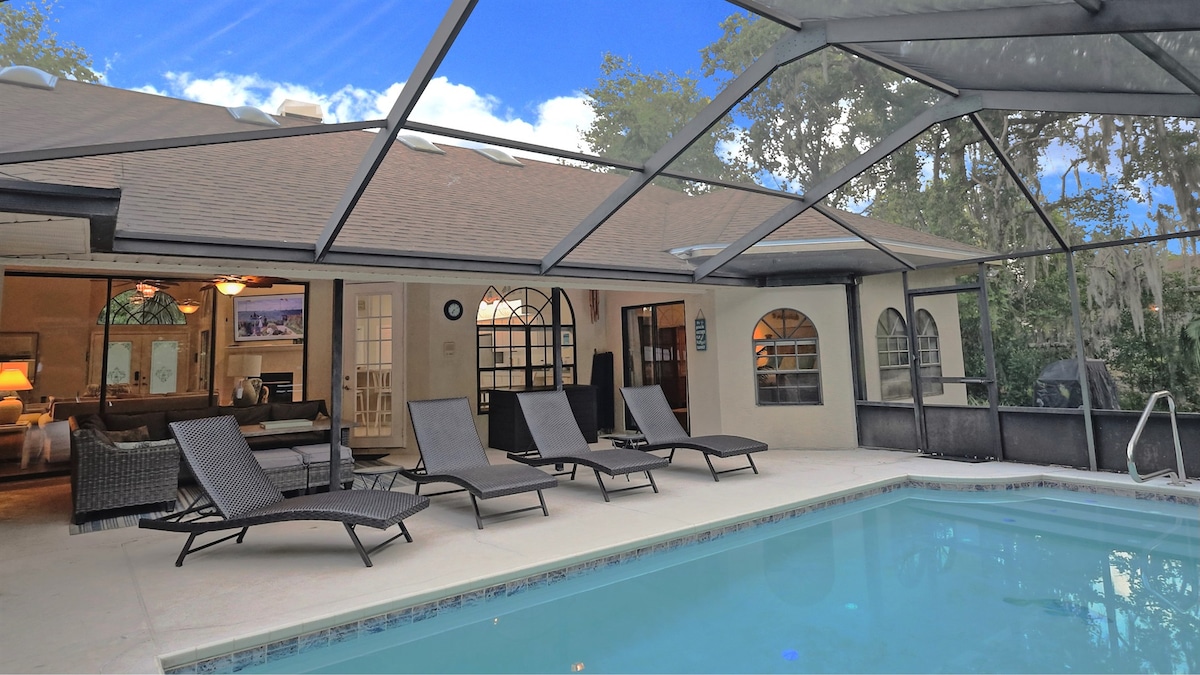 A screened lanai surrounds a heated pool, featuring four spacious, cushioned loungers arranged for relaxation. The expansive glass doors provide a view of the open living area, while the backdrop of mature trees offers privacy and a natural feel.
