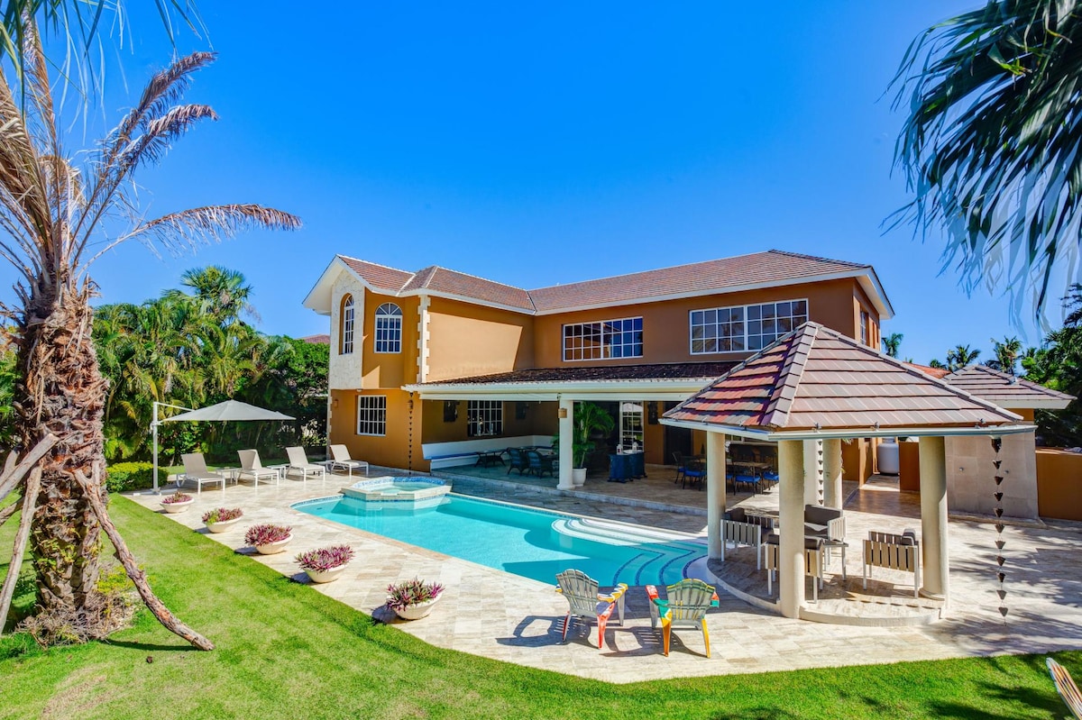 The villa's exterior showcases a spacious patio with a private swimming pool, surrounded by well-maintained landscaping. An outdoor gazebo provides shaded seating, while vibrant palm trees offer a tropical ambiance. Large windows allow natural light to illuminate the house's inviting façade.