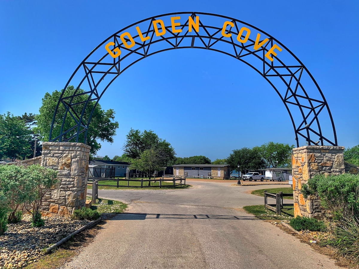 An entrance archway marked 'Golden Cove' stands prominently over a gravel road. Flanked by greenery, the road leads into a welcoming area with buildings visible in the distance under a clear blue sky.