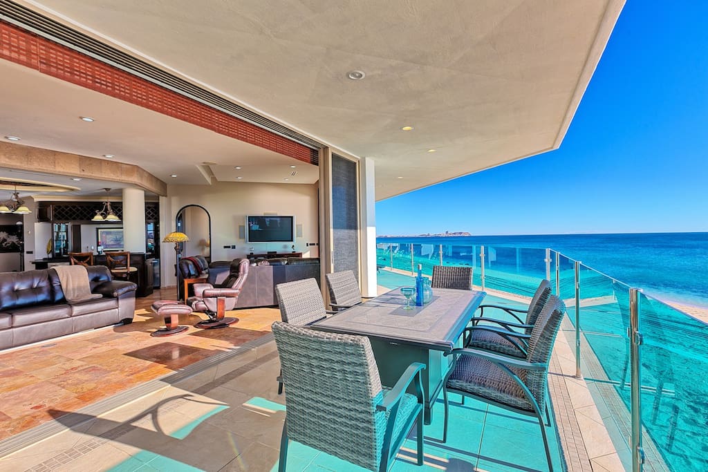 An expansive wrap-around patio is displayed, featuring cushioned outdoor chairs and a dining table set against stunning ocean views. The open layout connects to a spacious living area with large windows, allowing natural light to highlight the serene coastal atmosphere.