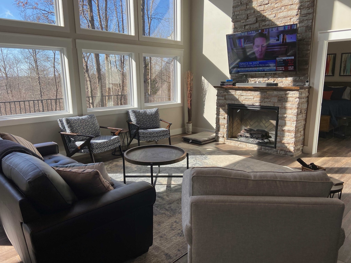 A bright living area features large windows, allowing natural light to fill the space. Comfortable seating includes a gray armchair and black sofas, arranged around a round table. A stone fireplace provides warmth, while a mounted television offers entertainment options.