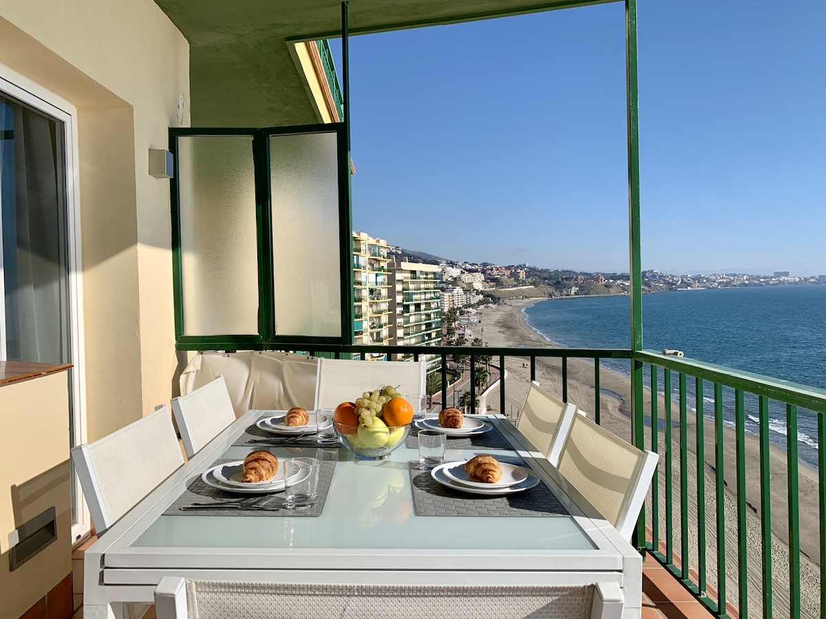 A spacious terrace is shown, featuring a glass dining table set for six, with plates containing croissants and fresh fruit. A clear view of the beach and sea is visible in the background, framed by green railings and the coastal landscape under a bright sky.
