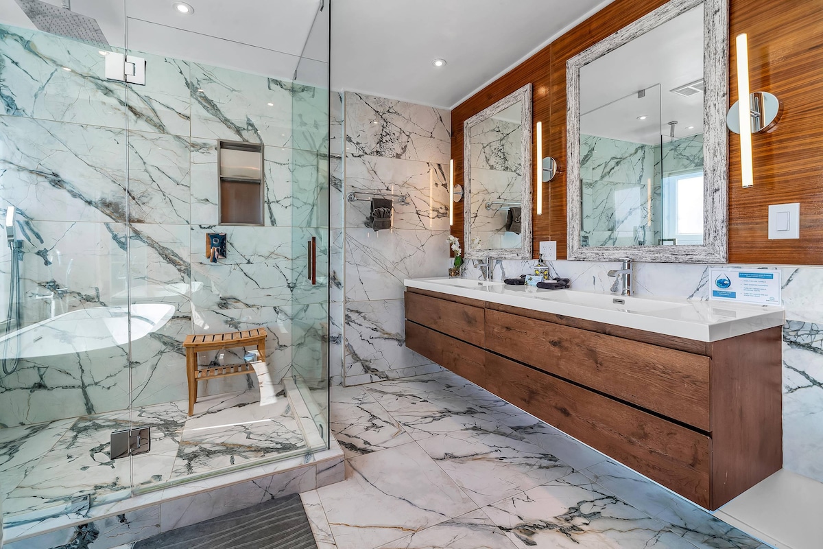 The modern bathroom features a glass-enclosed walk-in shower with a rainfall showerhead. A sleek double vanity with wooden cabinets is paired with large mirrors, complemented by marble walls. Natural light fills the space through a window, adding to the tranquil ambiance.